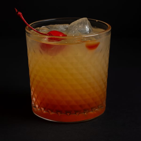 A vibrant orange and red cocktail with ice and a cherry in a textured glass.