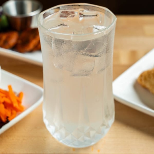 A tall glass of iced water on a wooden table, with Vietnamese dishes blurred in the background.