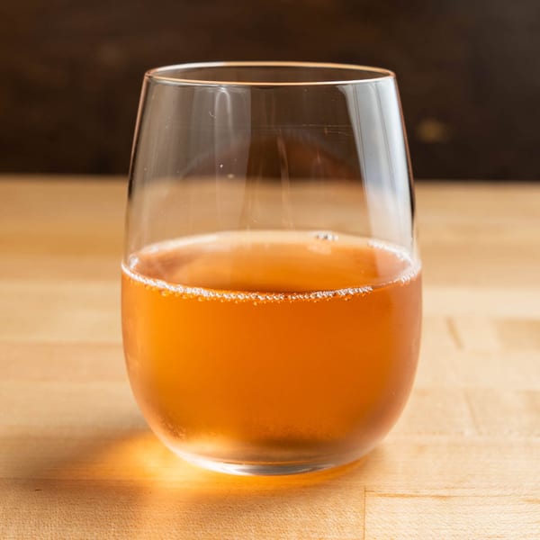 A stemless glass filled with a sparkling, amber-colored beverage on a light wooden surface.