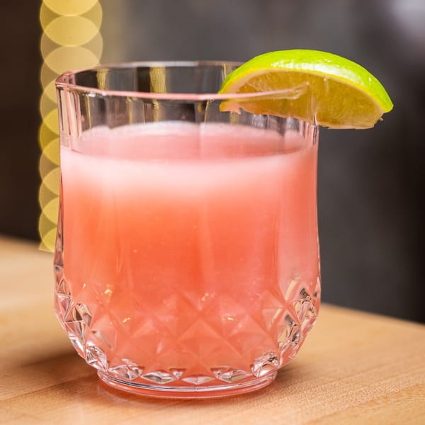 A vibrant pink cocktail in a textured glass, garnished with a fresh lime wedge, on a wooden surface.