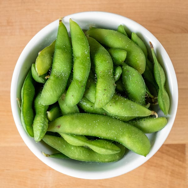 A white bowl filled with vibrant green edamame pods, lightly sprinkled with salt, on a wooden surface.